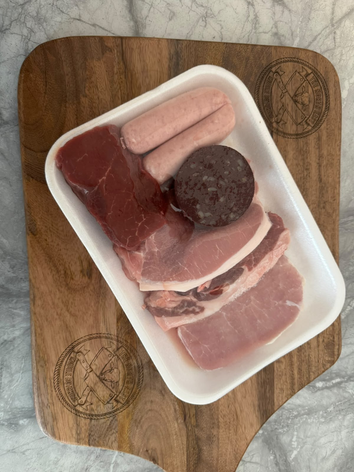 A tray of butcher-prepared steak, sausages, black pudding and bacon on an engraved North East Mobile Butchers board